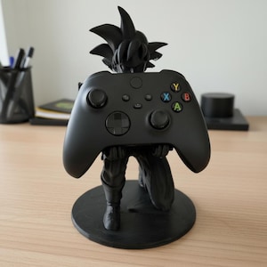 May include: A black video game controller held by a black figurine of a character with spiky hair. The controller has colored buttons and is resting on a black circular base. The figurine is kneeling.