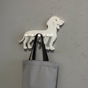 Könnte beinhalten: Ein weißer Dackel-Wandhaken mit drei Haken, der eine graue Tragetasche mit schwarzen Trägern hält. Der Haken ist an einer strukturierten grauen Wand angebracht und bietet eine dekorative und funktionale Aufbewahrungslösung.