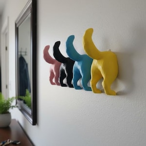 Może przedstawiać: Rząd pięciu kolorowych wieszaków ściennych w kształcie kocich tyłków. Wieszaki pomalowane są na różowo, czarno, niebiesko i żółto. Wieszaki zamontowane są na białej ścianie.