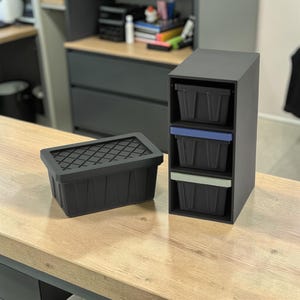 May include: A black storage bin with a cross-hatched lid sits next to a three-drawer storage unit. The drawers have blue and green accents. The items are on a light wood-grain surface.