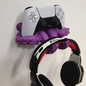 May include: A purple octopus-shaped wall mount holding a white and black game controller and a pair of black and red headphones. The controller is centered above the octopus, with the headphones hanging below.