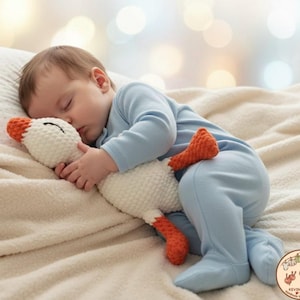 May include: A sleeping baby wearing a light blue onesie cuddles a white and orange crochet goose toy. The toy has a textured body and is being held close to the baby's face. The background is blurred with soft, warm lighting.