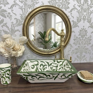 Pode incluir: Uma pia retangular de banheiro com um padrão floral verde e branco. A pia tem uma borda verde e é complementada por uma torneira dourada e um espelho redondo dourado. Uma xícara e saboneteira combinando também estão presentes.