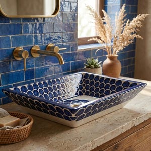 May include: A rectangular ceramic vessel sink featuring a hand-painted blue and white scalloped fish scale pattern. The basin has a glossy finish and is mounted on a textured stone countertop beneath a gold-toned faucet and blue tiled wall.
