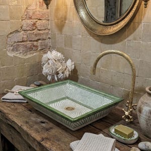 May include: A rectangular ceramic sink with a green and white geometric pattern. The sink sits on a wooden vanity with a gold faucet. A bar of soap rests on a small dish. A bouquet of white flowers and a mirror are in the background.