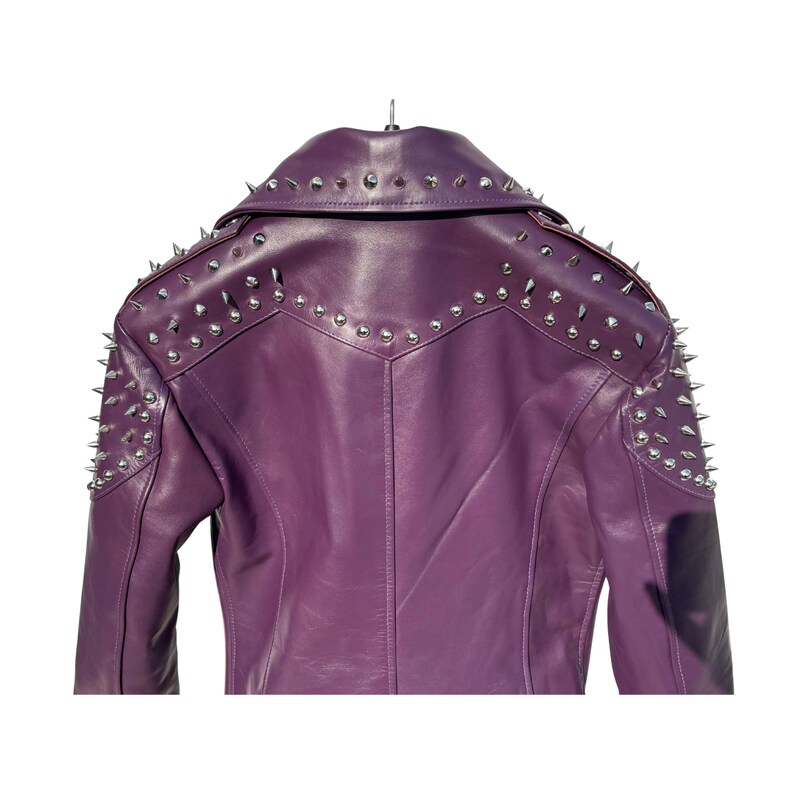 May include: A purple leather jacket with silver metal studs and spikes. The back of the jacket is visible, showing the collar, shoulders, and upper sleeves. The jacket is hanging on a hanger against a white background.
