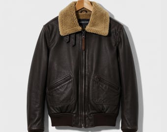 May include: A brown leather jacket with a tan shearling collar. The jacket has two front pockets, a zipper closure, and ribbed cuffs and hem. The jacket is hanging on a wooden hanger.