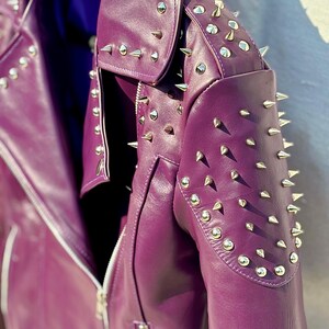 May include: A close-up of a purple leather jacket with silver metal cone studs. The jacket has a zipper closure and a notched collar. The shoulders and collar are heavily studded. The jacket is a vibrant shade of purple.