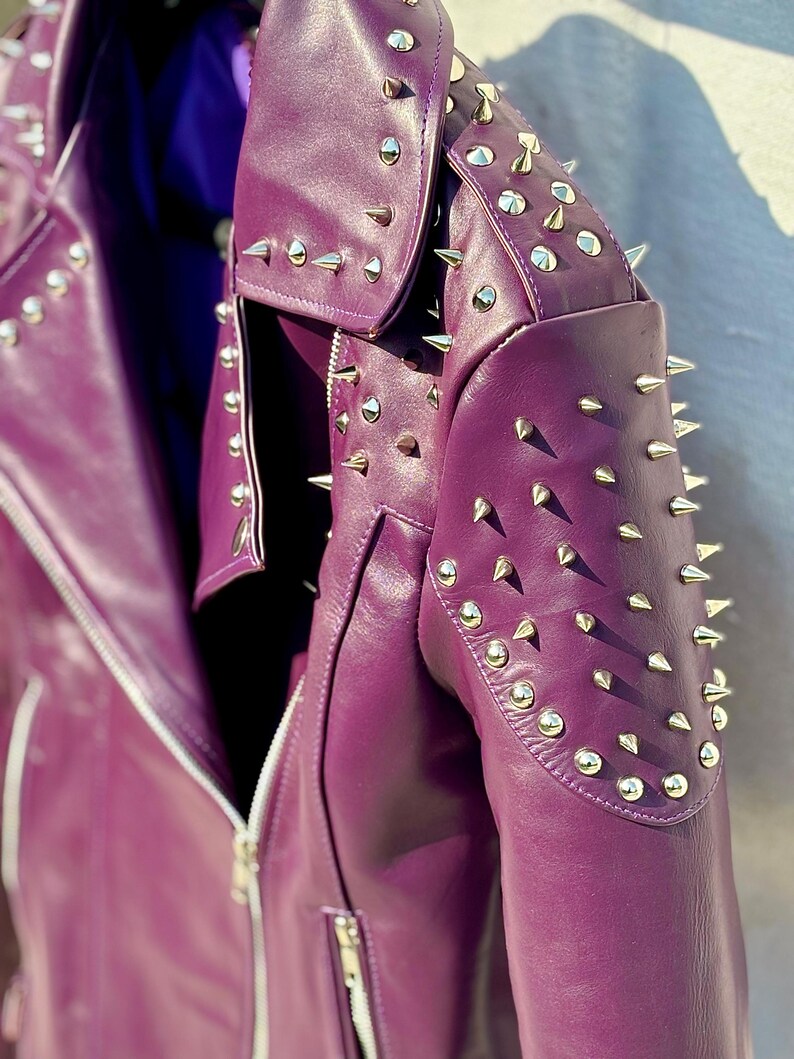 May include: A close-up of a purple leather jacket with silver metal cone studs. The jacket has a zipper closure and a notched collar. The shoulders and collar are heavily studded. The jacket is a vibrant shade of purple.