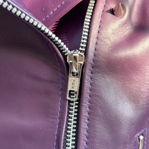 May include: Close-up of a purple leather jacket with a silver zipper and silver studs. The jacket has a shiny finish and a classic biker style. The zipper is marked with the letters "YKK".