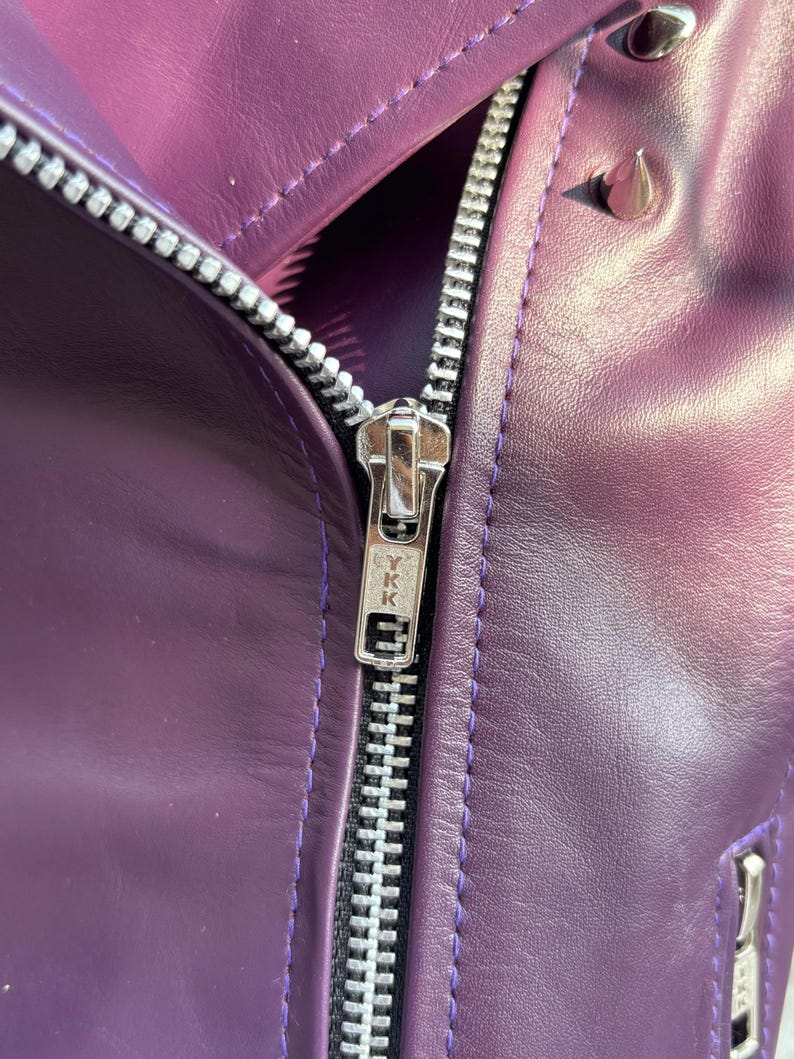 May include: Close-up of a purple leather jacket with a silver zipper and silver studs. The jacket has a shiny finish and a classic biker style. The zipper is marked with the letters "YKK".