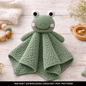 May include: A handmade green crochet frog security blanket with a smiling face, black eyes, and pink cheeks. The blanket has a square shape with a textured pattern. The image also includes a wooden rattle, a wooden bead garland, and star-shaped decorations.
