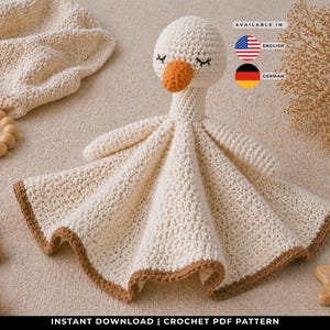 May include: A cream-coloured crochet baby blanket shaped like a goose, with an orange beak and closed eyes. The blanket has a brown border. Wooden toys and a wooden teething ring are also in the image. The text "INSTANT DOWNLOAD | CROCHET PDF PATTERN" is at the bottom.