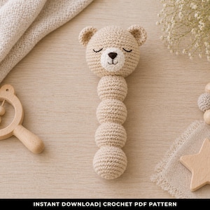 May include: A beige crochet bear rattle with a smiling face and a series of stacked spheres. A wooden rattle and a star-shaped teether are also present. The image is of baby toys.
