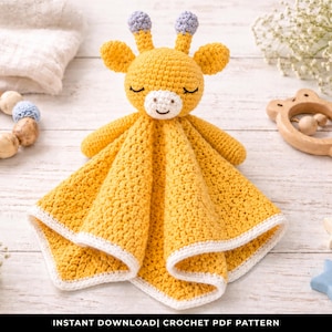 May include: A handmade yellow crochet giraffe security blanket with a white border. The giraffe has closed eyes, small ears, and purple accents. A wooden rattle, wooden beads, and star-shaped toys are also visible.
