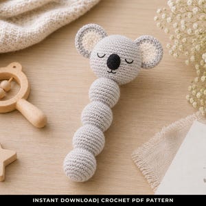 May include: A handmade crochet koala rattle in light gray with a black nose and closed eyes. A wooden star and rattle are next to the koala. The text "for you" is on a card.