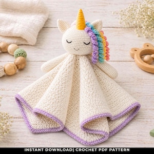 May include: A cream-colored crochet unicorn security blanket with a purple border. The unicorn has a yellow horn, rainbow mane, and closed eyes. Other items include a wooden rattle, wooden beads, and star-shaped decorations. The text at the bottom reads "INSTANT DOWNLOAD| CROCHET PDF PATTERN".