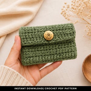 May include: A handmade, olive green crochet wallet with a wooden button closure. The wallet is rectangular with a flap and is held in a hand. The button has a floral design. The text "INSTANT DOWNLOAD| CROCHET PDF PATTERN" is at the bottom.