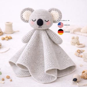 May include: A handmade crochet koala security blanket in light gray. The koala has a black nose, pink cheeks, and closed eyes. The blanket is surrounded by other baby toys, including a wooden ring, a wooden teether, and a stackable wooden toy.