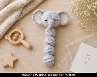 May include: A gray crochet elephant rattle with a long body of connected spheres. The elephant has closed eyes and large ears with a cream-colored interior. A wooden star and rattle are also present, along with a card that says "for you."