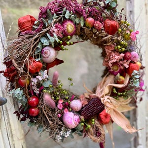 Könnte beinhalten: Ein runder Kranz aus braunen Zweigen, verziert mit getrockneten rosa Blumen, roten Physalis-Hüllen, kleinen roten Äpfeln, grünen Beeren, hellrosa Schneckenhäusern, violettem Mais und getrockneten Eukalyptusblättern, aufgehängt an einem dicken Juteseil.