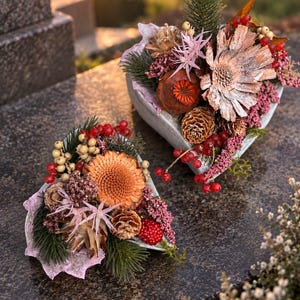 Könnte beinhalten: Zwei herzförmige Blumenarrangements mit getrockneten orangefarbenen Blüten, Tannenzapfen, grünen Kiefernnadeln, roten Beeren und hellrosa Akzenten. Diese dekorativen Stücke bestehen aus natürlichen botanischen Elementen in dichter Anordnung.