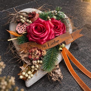 Könnte beinhalten: Eine herzförmige Steinplatte, verziert mit zwei dunkelroten Rosen, getrockneten Lotusblüten, Tannenzweigen und kleinen weißen Beeren. Ein rostiges Metallband in der Mitte trägt die Gravur In Liebe, ergänzt durch zwei lange orangefarbene Bänder.