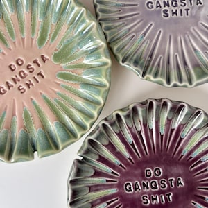 May include: Three round ceramic dishes with scalloped, sunburst-patterned edges. One dish is pink with green accents, one is light purple, and one is dark purple. Each dish features the embossed text DO GANGSTA SHIT in the center.