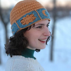 May include: A handmade crochet hat in tan and blue. The hat has a blue band with decorative tan shapes. The hat looks warm and comfortable. A stylish accessory for cooler weather. The hat is a unique item.