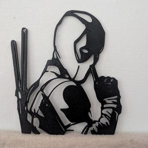 Könnte beinhalten: Ein Wandkunstobjekt aus schwarzem Metall, das die stilisierte Silhouette einer maskierten Figur mit Kapuze zeigt. Die Figur trägt Handschuhe und zwei lange, schmale, schwertähnliche Waffen auf dem Rücken. Das Design ist aus Metall geschnitten.