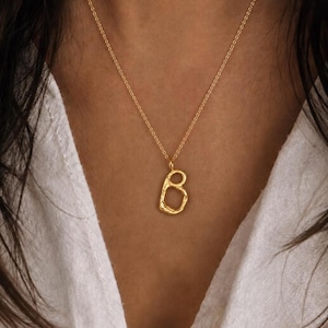 Pode incluir: Um colar dourado com um pingente em forma da letra "B". O pingente texturizado pende de uma corrente fina. O colar é usado sobre uma camisa branca.