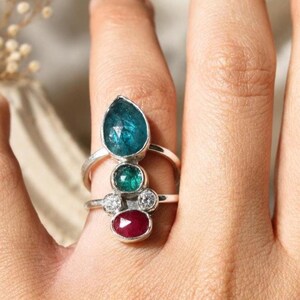 Könnte beinhalten: Drei Edelsteinringe an einem Finger. Der obere Ring hat einen großen, tropfenförmigen tealfarbenen Stein. Der mittlere Ring hat einen kleinen, runden Smaragd. Der untere Ring hat einen Rubin und zwei kleine weiße Steine. Alle Ringe haben silberne Bänder.