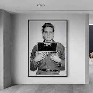 May include: Black and white framed print of Elvis Presley holding a sign that reads "Presley Elvis A". He is wearing a military uniform. The image is displayed on a white wall in a modern hallway.