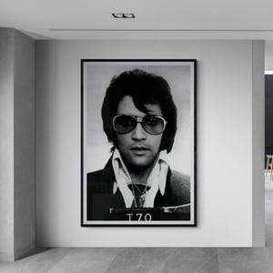 May include: Black and white framed portrait of a man wearing sunglasses, a collared shirt, and a jacket. The image is a mugshot, with the number "T70" visible at the bottom. The portrait is displayed on a white wall in a modern interior.