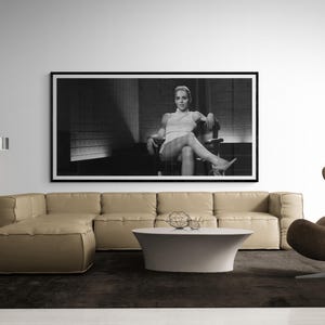 May include: Black and white framed photograph of a woman seated in a chair. The photograph is displayed above a beige sectional sofa, with a white coffee table and a dark brown rug in the foreground. The room has a modern, minimalist design.