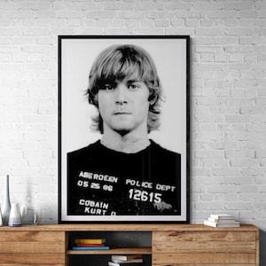 May include: Black and white framed mugshot of a man with long, light-colored hair. The man is wearing a black shirt with text that reads "Aberdeen Police Dept 12615 Cobain Kurt D 05 25 86". The image is displayed on a wooden cabinet.