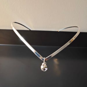 Puede incluir: Un collar gargantilla plateado con un diseño minimalista. El collar presenta una fina banda en forma de V. Un pequeño colgante redondo de piedra preciosa transparente cuelga del centro de la V, añadiendo un toque de brillo.