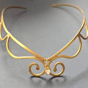 May include: A gold-colored metal tiara with a delicate design. The tiara features curved lines and a central pendant with a small, clear gemstone. The overall design is elegant and ornate, suitable for special occasions.