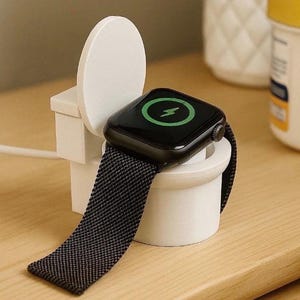 Könnte beinhalten: Eine schwarze Apple Watch mit schwarzem Mesh-Armband liegt auf einer weißen Ladestation. Das Zifferblatt zeigt einen grünen Blitz, der das Laden anzeigt. Der Ständer hat die Form einer Miniaturtoilette.
