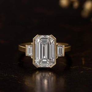 May include: A gold ring with a large, rectangular, emerald-cut diamond as the centerpiece, flanked by two smaller, rectangular diamonds. The ring is set against a dark background, highlighting the sparkle of the diamonds.