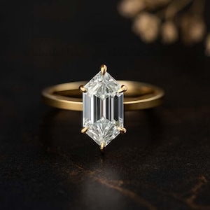 Puede incluir: Un anillo de oro con un diamante de talla hexagonal. El diamante claro y facetado refleja la luz. La delgada banda de oro del anillo contrasta con un fondo oscuro. El anillo es una joya.