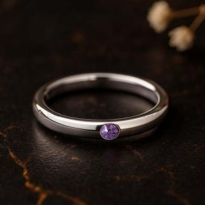 Puede incluir: Un anillo de plata pulida con una pequeña gema redonda de color púrpura. El anillo tiene un acabado liso y se encuentra sobre una superficie oscura y texturizada. Una joya sencilla y elegante.
