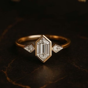May include: A gold ring featuring a central emerald-cut diamond, with two smaller diamond accents. The ring has a geometric design. The gemstones sparkle against a dark background, highlighting the ring's elegant design.
