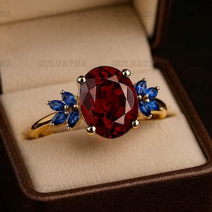 Puede incluir: Un anillo de oro con una gran piedra preciosa roja oscura de talla ovalada como pieza central. El anillo está adornado con grupos de pequeñas piedras preciosas azules a ambos lados de la piedra principal. El anillo se presenta en un joyero marrón.