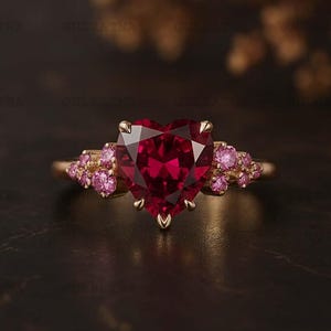 Op de afbeelding: Een gouden ring met een hartvormige, gefacetteerde robijn als centrale steen. Kleinere roze edelstenen flankeren de robijn. De ring is geplaatst tegen een donkere achtergrond.