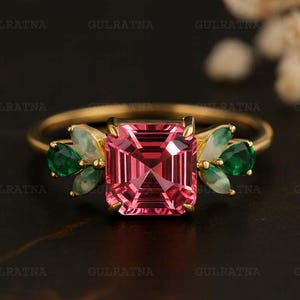 Puede incluir: Un anillo de oro con una gran piedra rosa de corte cuadrado en el centro. El anillo está adornado con piedras preciosas verdes y blancas más pequeñas. El anillo está sobre un fondo oscuro.