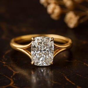 Puede incluir: Un anillo de compromiso dorado con una gema clara grande y rectangular. El anillo tiene un diseño de caña dividida, con la banda dividiéndose en dos curvas delicadas que acunan la gema. El anillo está sobre un fondo oscuro y texturizado.