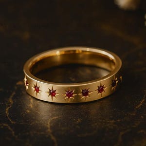 May include: A gold-colored ring with a series of star-shaped cutouts, each containing a small, red gemstone. The ring is set against a dark, textured background, highlighting the ring's design and color.