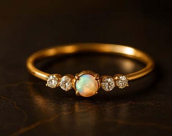 Puede incluir: Un delicado anillo de oro con una piedra central de ópalo flanqueada por tres diamantes más pequeños y brillantes a cada lado. El anillo tiene una banda fina y está engastado sobre un fondo oscuro, resaltando las piedras preciosas.