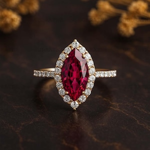 Op de afbeelding: Een gouden ring met een marquise-geslepen, dieprode edelsteen, omringd door kleine, ronde diamanten. De band is ook bezet met kleine diamanten. De ring wordt getoond tegen een donkere, getextureerde achtergrond.